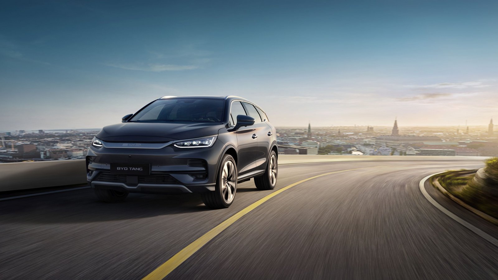 BYD Tang SUV fährt auf einer Landstraße mit Stadt im Hintergrund bei Sonnenuntergang