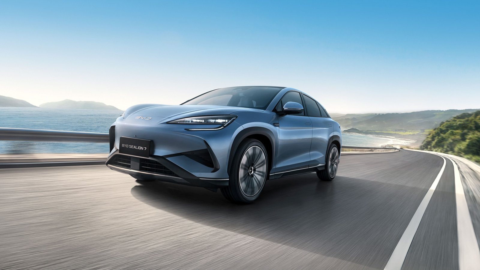 BYD SEALION Elektro-SUV in Blau fährt auf kurviger Landstraße mit Meer und Hügeln im Hintergrund